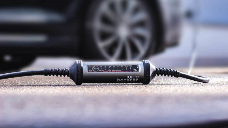 Nahaufnahme der robusten mobilen Ladestation Juice Booster im Außeneinsatz auf Asphalt mit einem unscharfen Fahrzeugrad im Hintergrund.